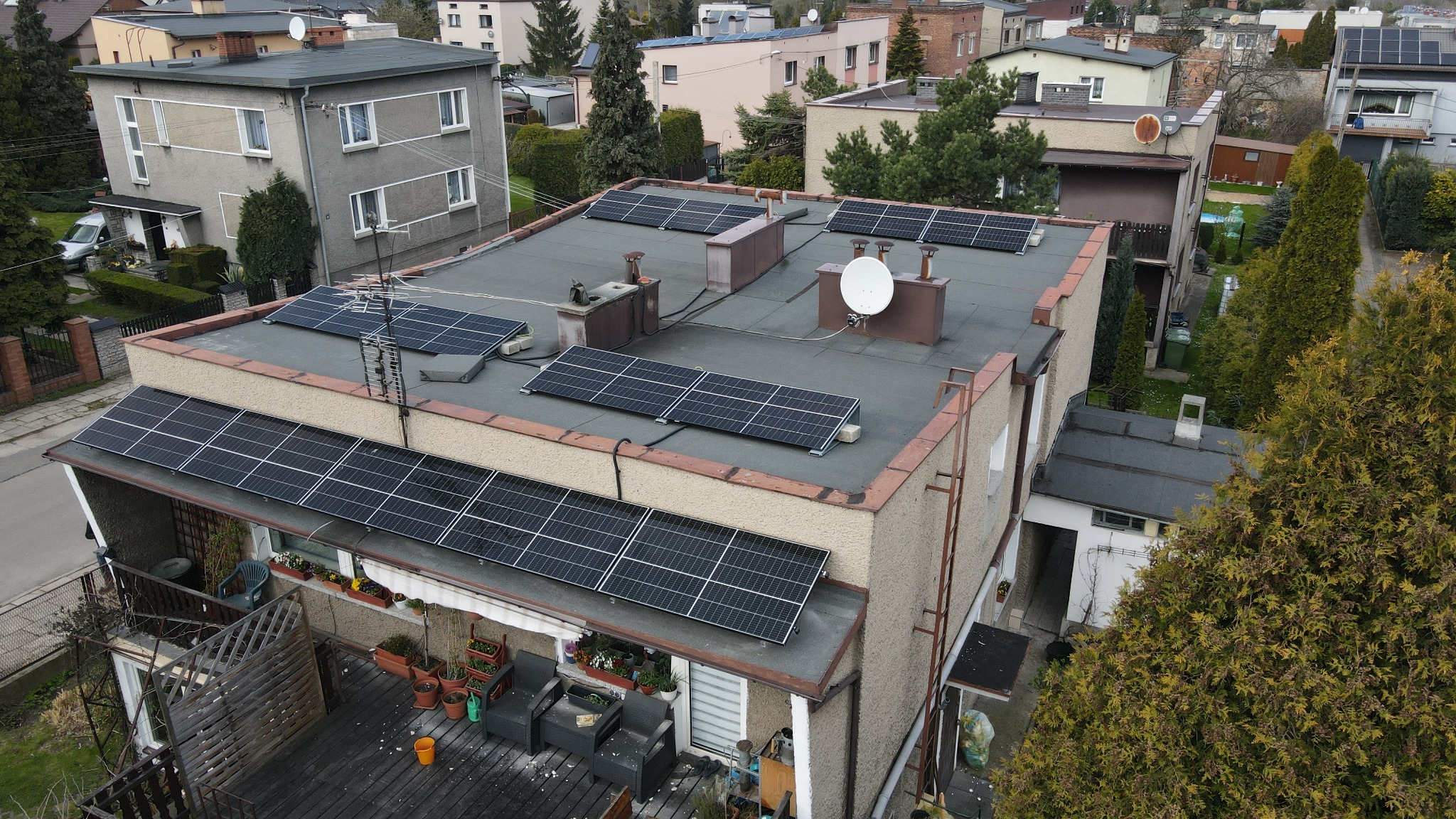 Widok z góry na dom z panelami słonecznymi na dachu i nad balkonem, w tle zabudowa osiedlowa.