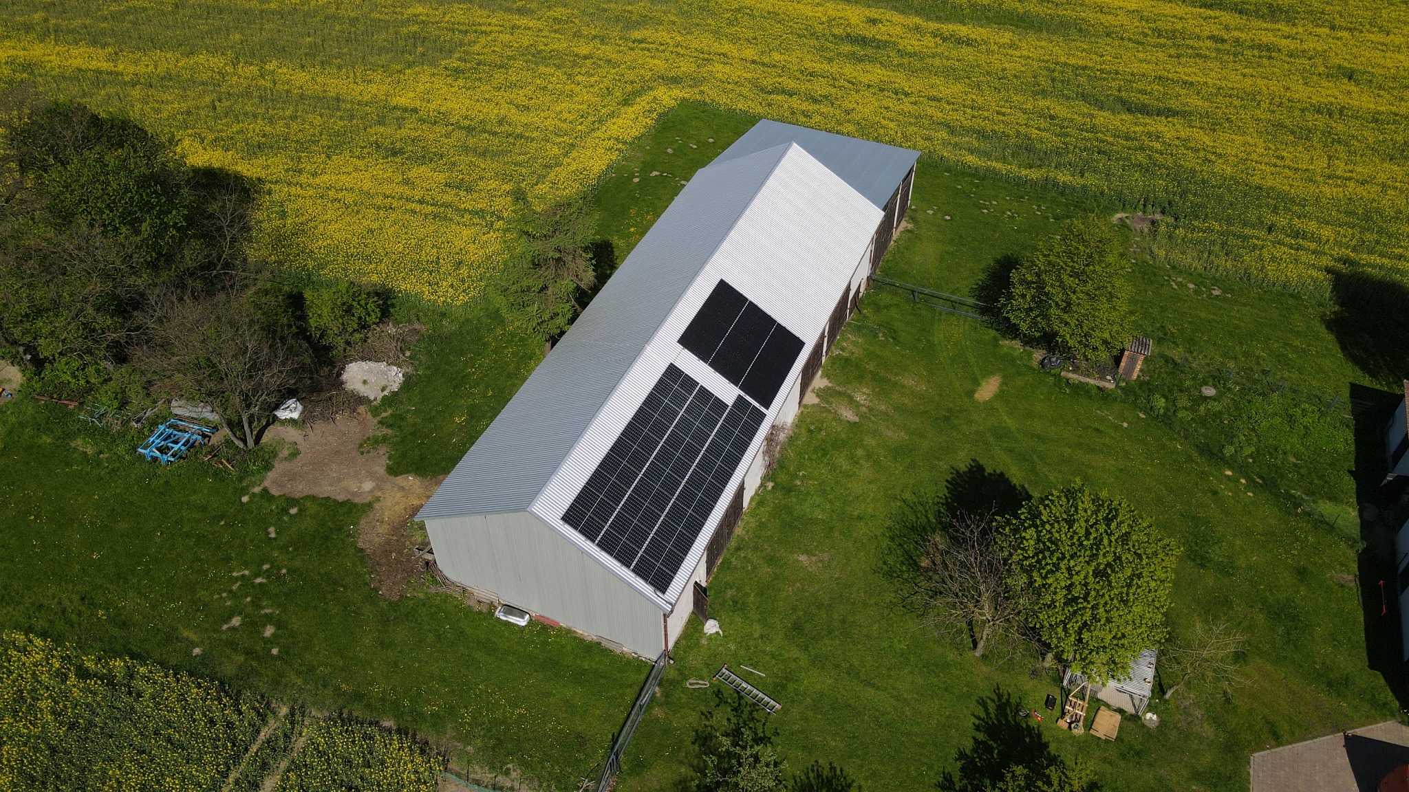 Widok z góry na metalowy budynek gospodarczy z panelami słonecznymi na dachu, otoczony zieloną łąką i kwitnącym polem rzepaku.