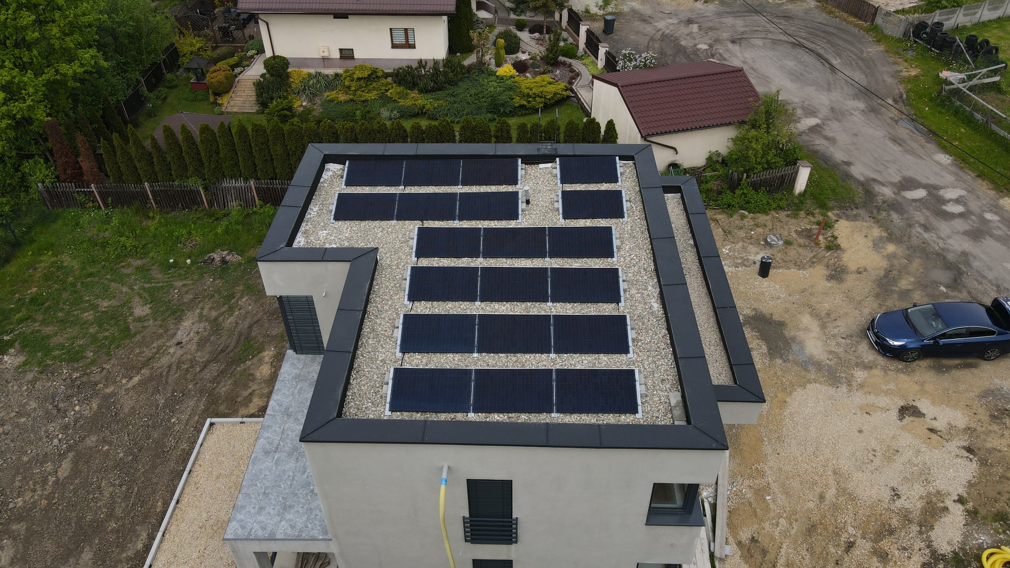 Widok z góry na nowoczesny dom z panelami słonecznymi zamontowanymi na płaskim dachu pokrytym drobnym żwirem, w tle zabudowa jednorodzinna i podjazd z zaparkowanym niebieskim samochodem.