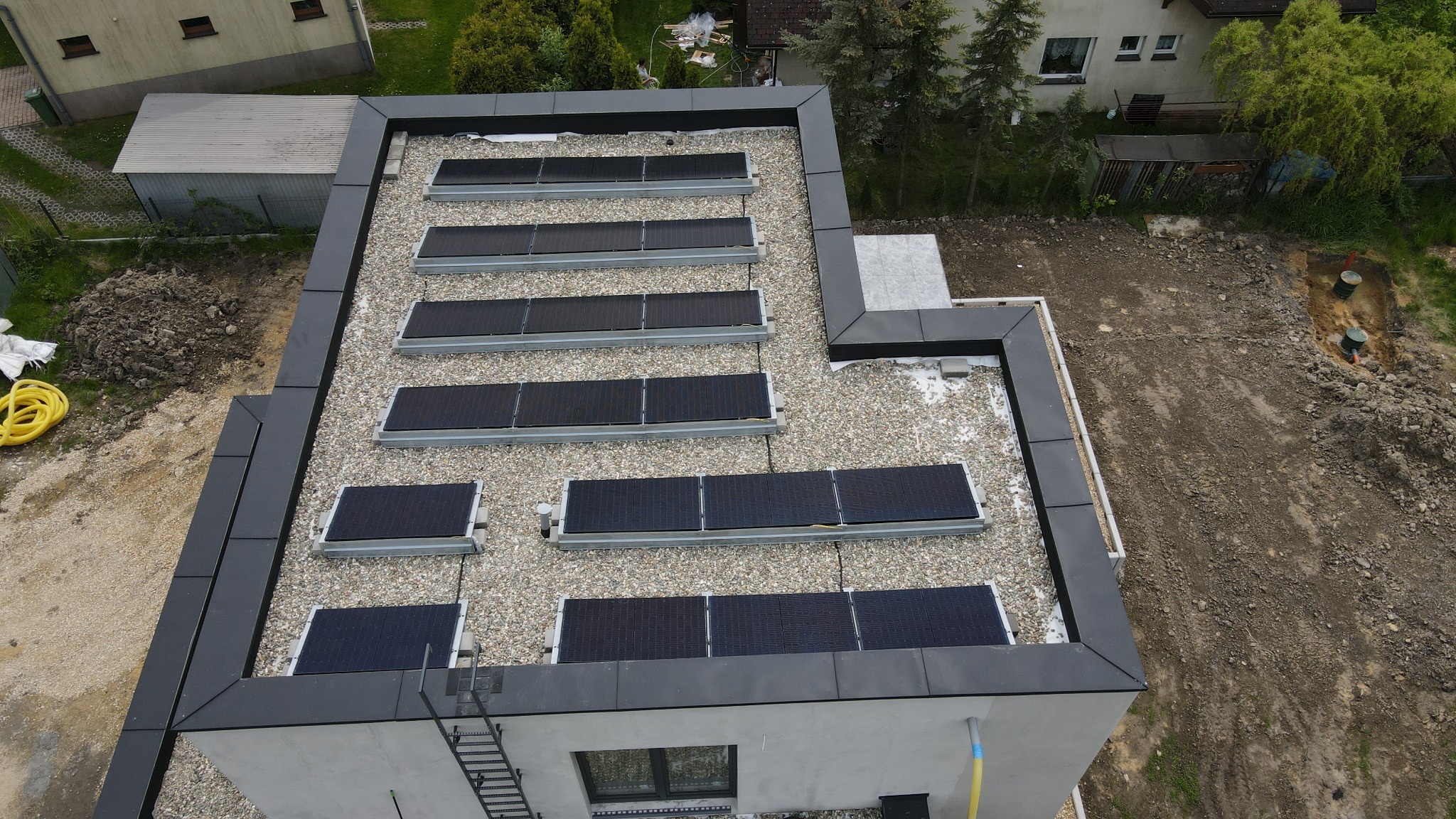 Widok z góry na płaski dach z panelami słonecznymi, pokryty drobnym żwirem, z fragmentem elewacji budynku i otoczeniem w tle.