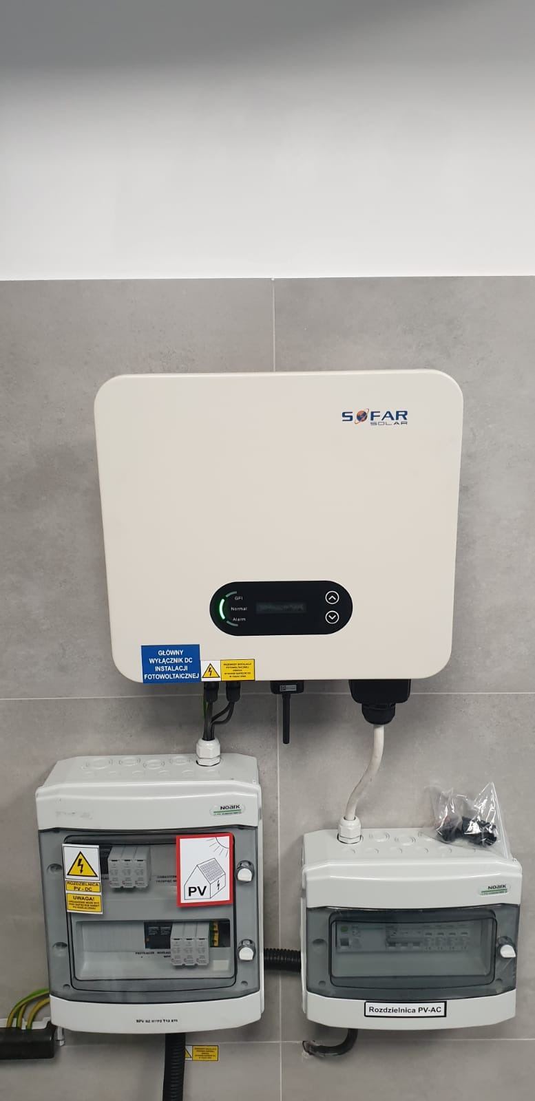 Biały inwerter Sofar Solar zamontowany na szarej ścianie kafelkowej, poniżej dwie szare skrzynki rozdzielcze PV-DC i PV-AC, widoczne kable i naklejki ostrzegawcze.