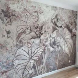 Ściana z fototapetą przedstawiającą abstrakcyjne kwiaty i liście w odcieniach szarości i brązu, widoczny fragment podłogi z paneli w ciepłym odcieniu drewna, biała listwa przypodłogowa i gniazdko...