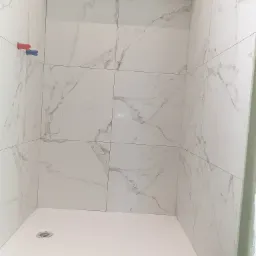 Narożne ujęcie kabiny prysznicowej z białym brodzikiem i ścianami wyłożonymi białymi płytkami imitującymi marmur, widoczne wystające rurki instalacji wodnej i fragment nieotynkowanej ściany.