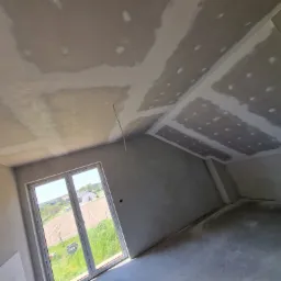 Wnętrze poddasza w trakcie prac wykończeniowych: sufit z płyt kartonowo-gipsowych z widocznymi łączeniami szpachlowanymi, ściany przygotowane pod malowanie, okno z widokiem na zielone pole...