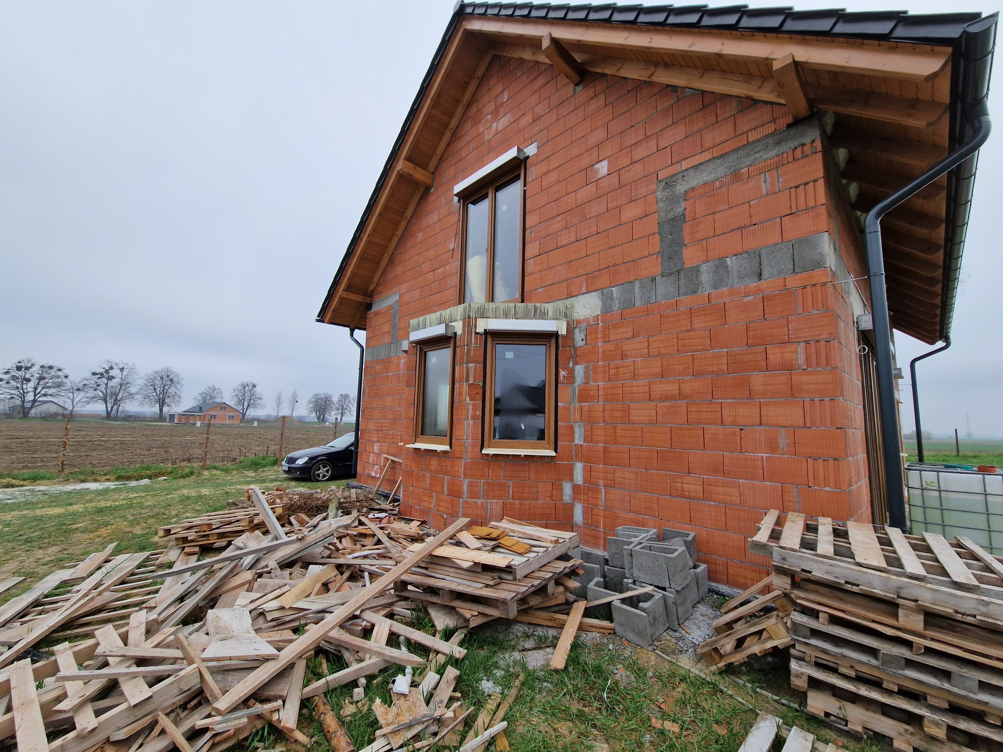 Surowy stan budynku z czerwonej cegły z widocznymi oknami, paletami i gruzem na trawniku, na tle pochmurnego nieba i pola z drzewami w oddali.