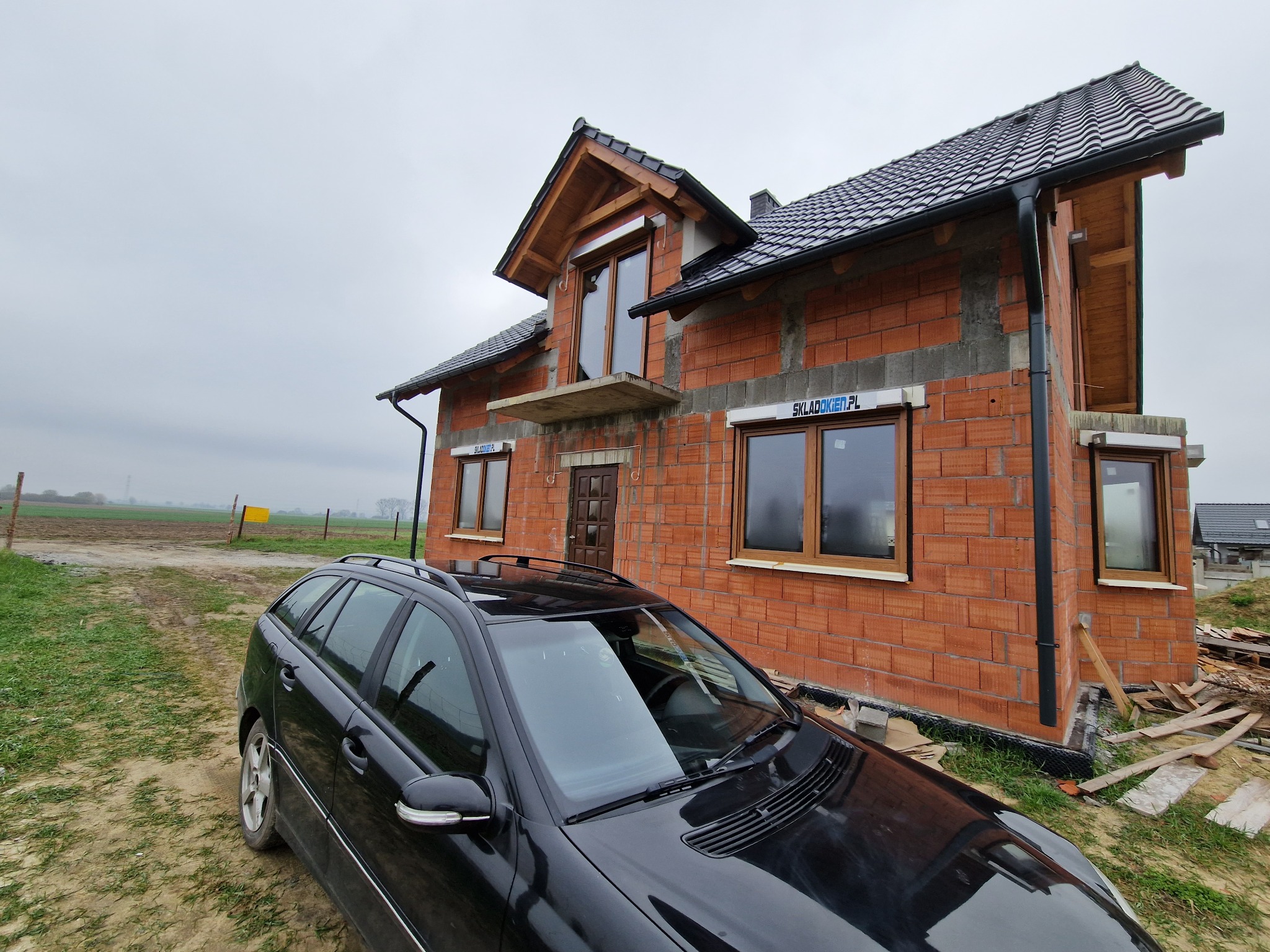 Surowy stan domu jednorodzinnego z czerwonej cegły z zamontowanymi oknami i dachem z dachówki, widok z perspektywy samochodu osobowego na pierwszym planie, pochmurne niebo w tle.