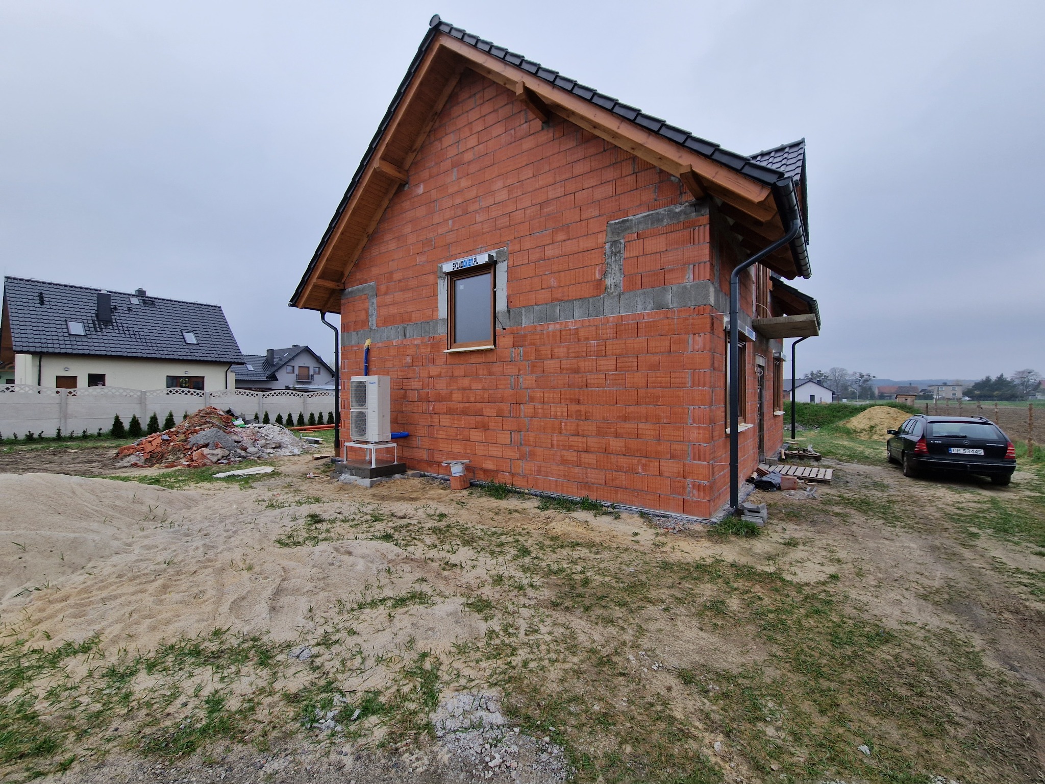 Niedokończony dom jednorodzinny z czerwonej cegły, widoczny klimatyzator zewnętrzny, okno z roletą i sterta gruzu na posesji.