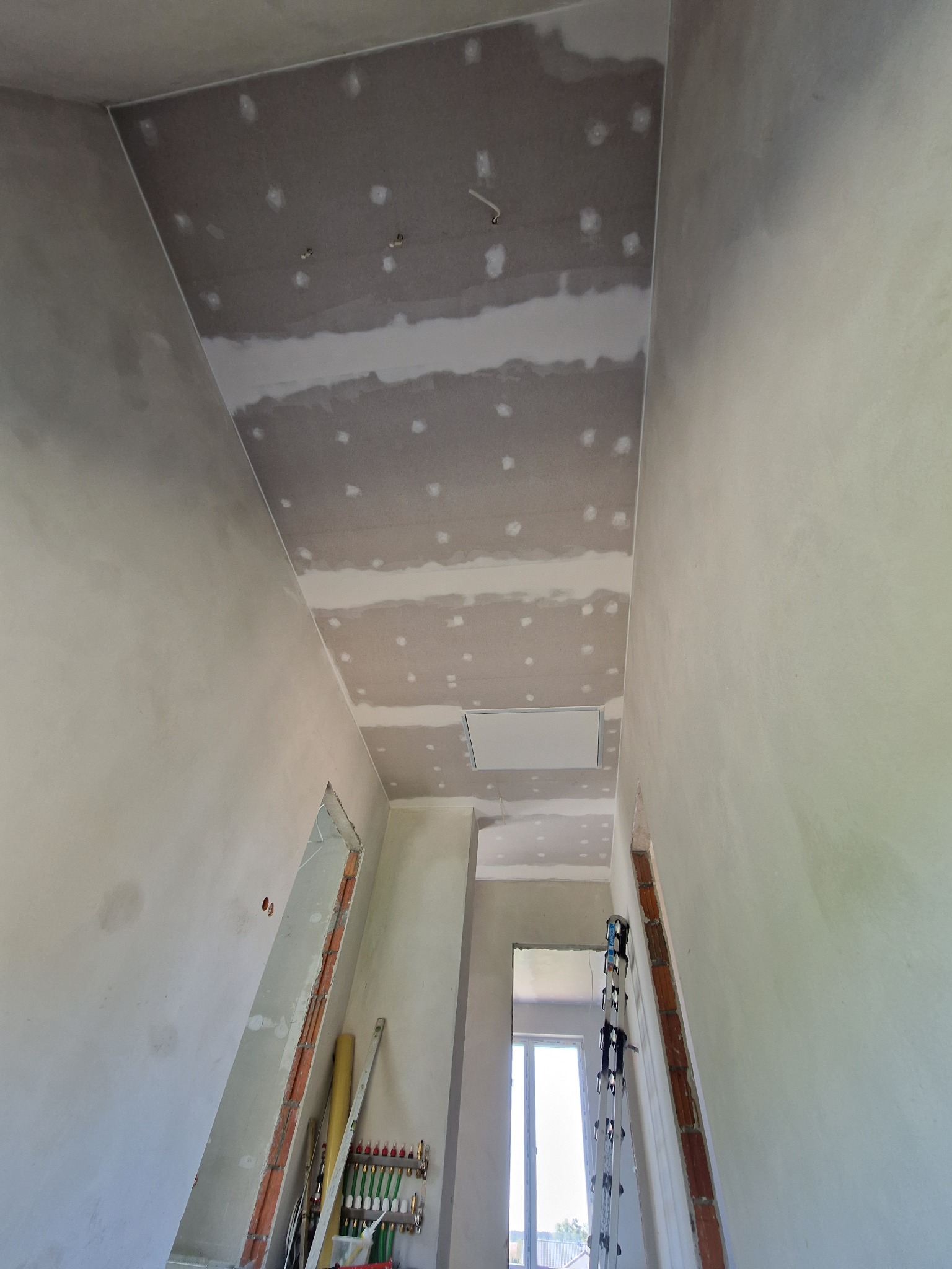 Wnętrze w trakcie prac wykończeniowych: ściany i sufit pokryte szpachlą, widoczne łączenia płyt gipsowo-kartonowych, instalacje grzewcze przy ścianie, drabina oparta o ścianę.