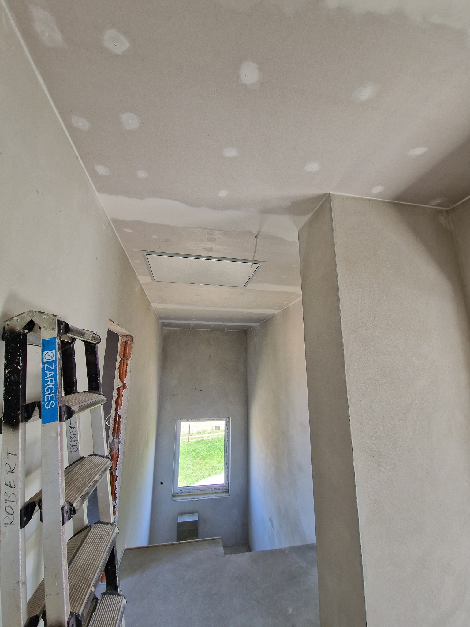 Wnętrze w trakcie prac w Opolu: widoczne ściany i sufit po szpachlowaniu płyt kartonowo-gipsowych, z widocznymi miejscami po wkrętach, aluminiowa drabina Zarges oraz okno w oddali.