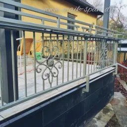 Szara, metalowa balustrada z ozdobnymi elementami kwiatowymi na tarasie wyłożonym jasnymi płytkami, z widokiem na ogród i dom w tle.