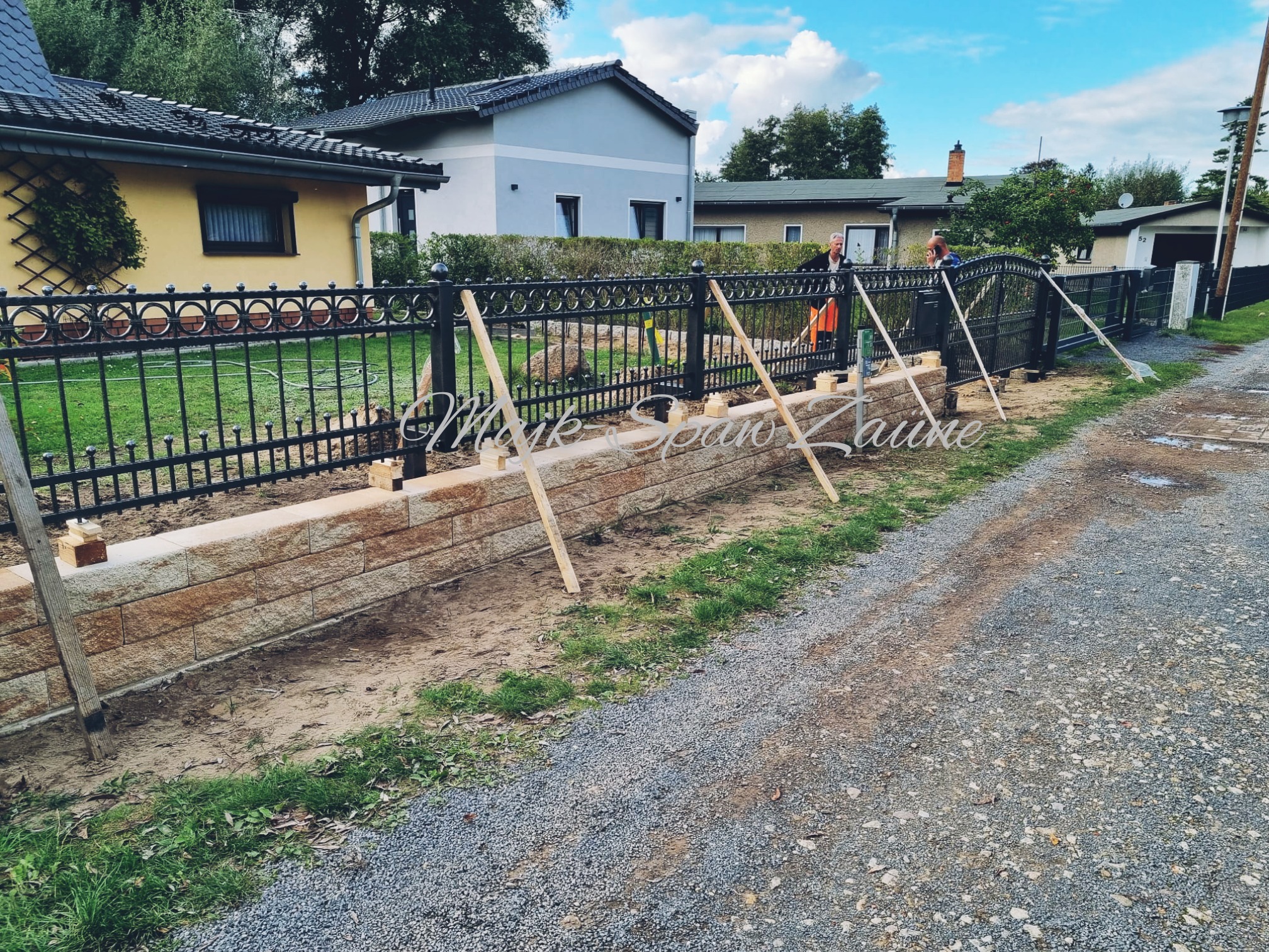 Montaż czarnego, metalowego ogrodzenia z ozdobnymi elementami nad niskim murem z kamienia piaskowca, widoczni dwaj pracownicy podczas instalacji, drewniane podpory stabilizują konstrukcję.
