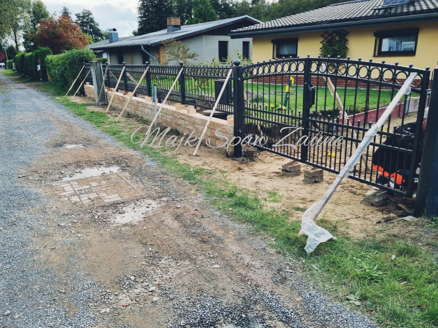 Montaż kutej bramy wjazdowej z ozdobnymi elementami, widoczne podpory tymczasowe i niezakończone prace ziemne.
