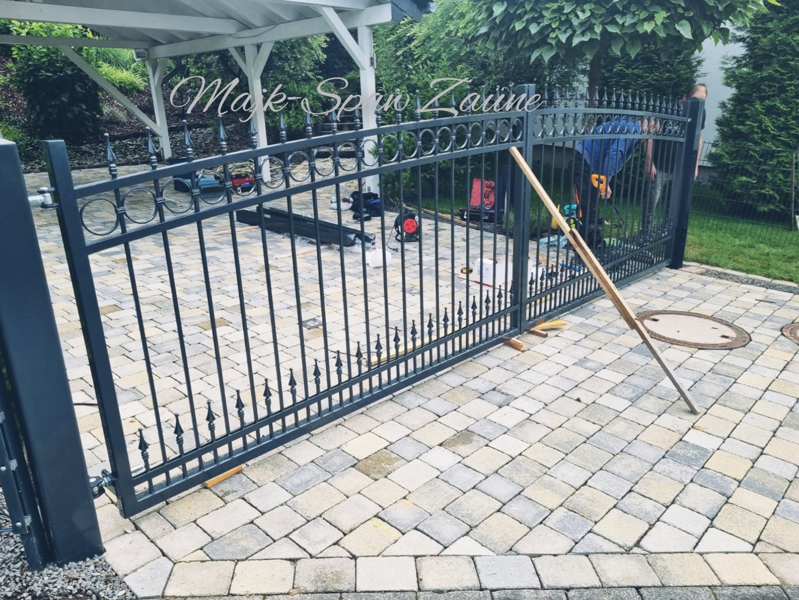 Ciemnoszara, metalowa brama z dekoracyjnymi elementami w trakcie montażu na posesji z kostki brukowej; widoczne narzędzia, deski podpierające i osoba w tle.