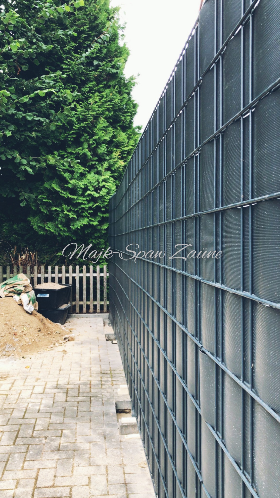 Montaż ogrodzenia panelowego z widoczną konstrukcją, częściowo zasłoniętą przez gęste drzewo iglaste, z workami z materiałem budowlanym i wiaderkiem w tle.