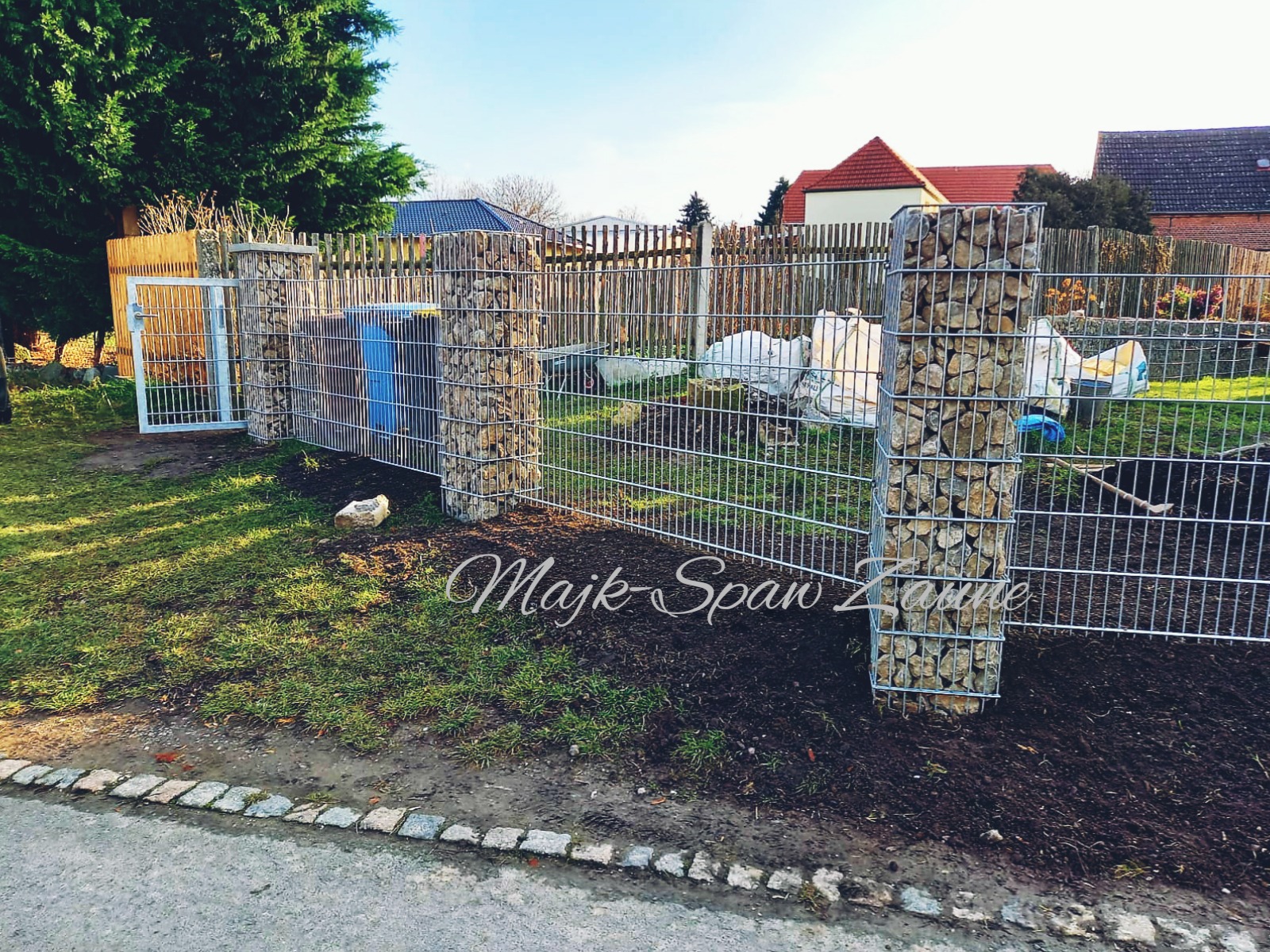 Ogrodzenie z metalowej siatki z kamiennymi słupkami, widoczna furtka, trawnik i zabudowa mieszkalna w tle.