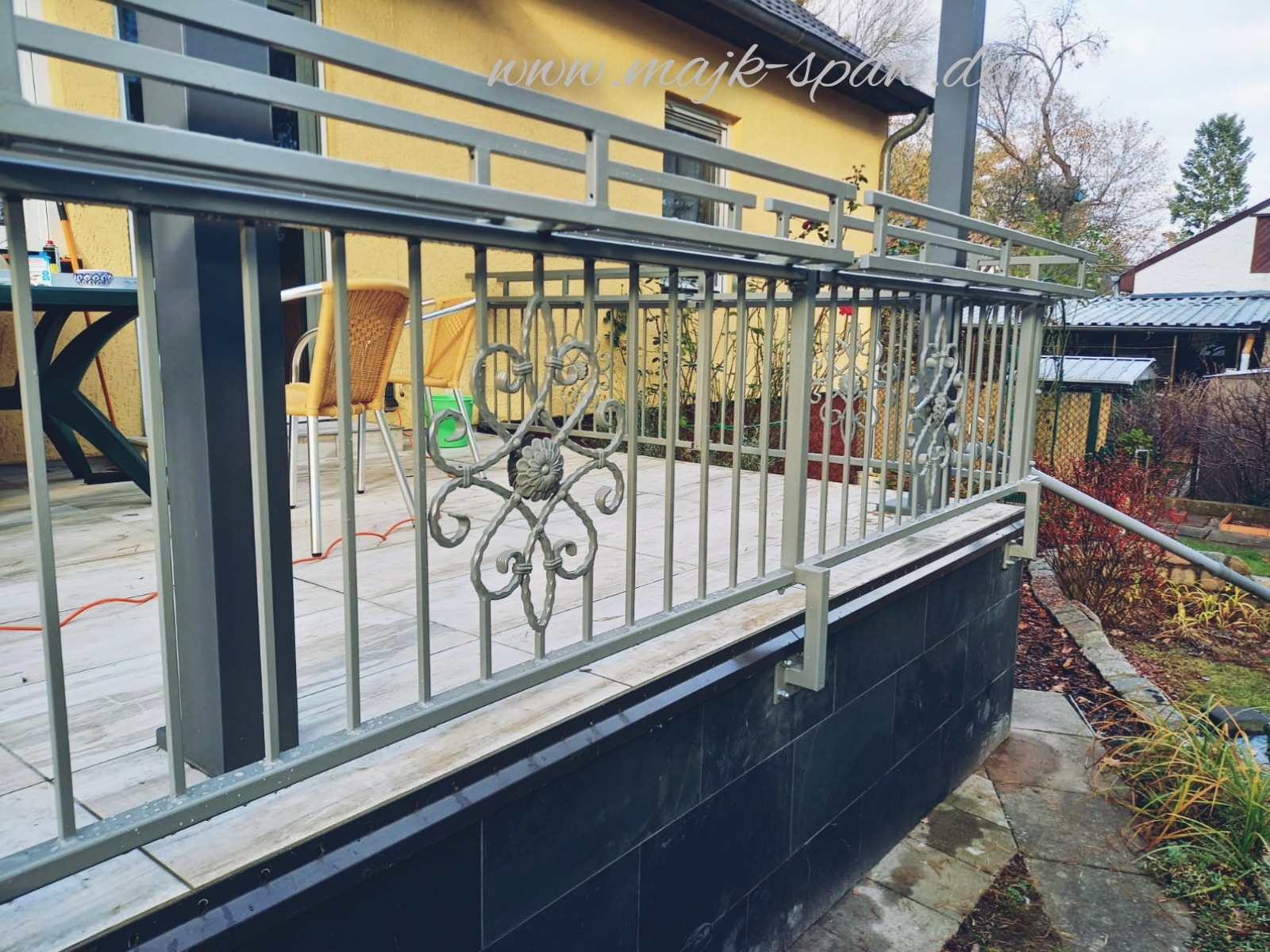 Szara, metalowa balustrada z ozdobnymi elementami kwiatowymi na tarasie wyłożonym jasnymi płytkami, z widokiem na ogród i dom w tle.