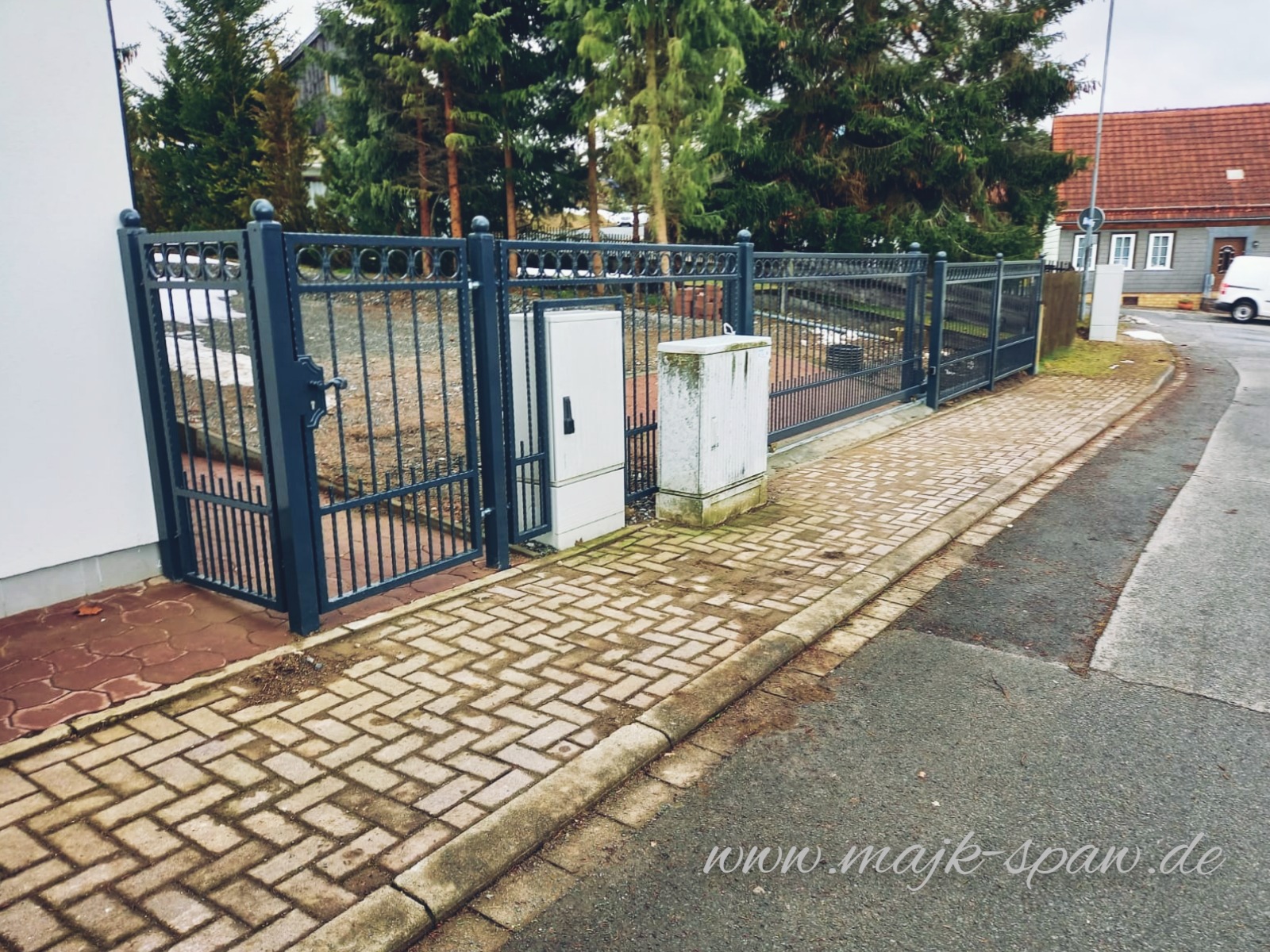 Ciemnoszara metalowa brama i ogrodzenie z prętów, z kulistymi zakończeniami, z widocznymi skrzynkami elektrycznymi obok, ułożone na kostce brukowej.