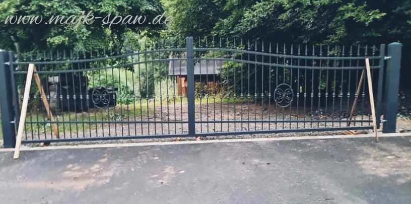 Szara, metalowa brama wjazdowa z dekoracyjnymi elementami, podparta drewnianymi listwami podczas montażu, widok na zieleń w tle.