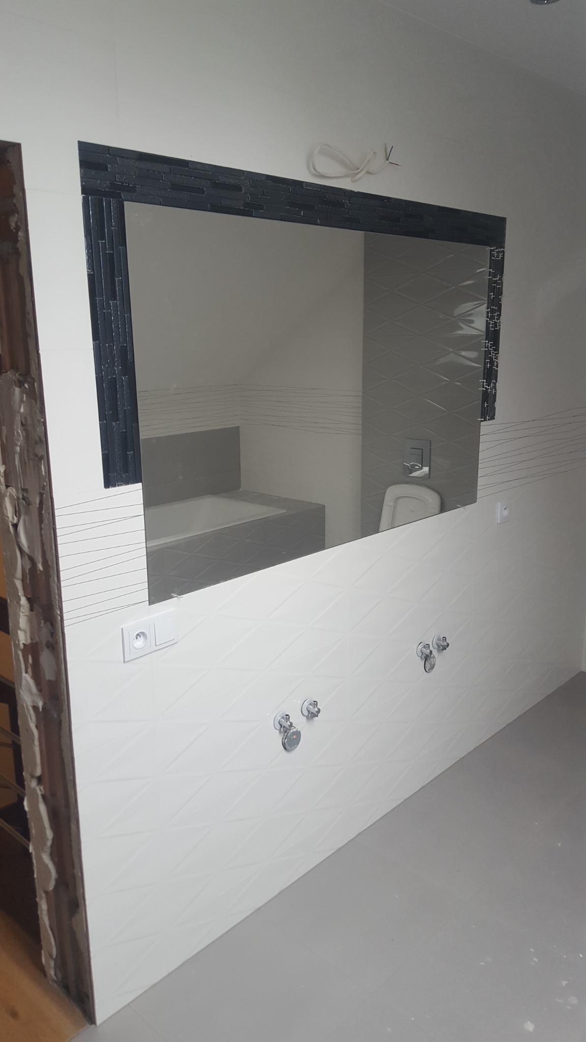 Nowoczesna łazienka w trakcie wykańczania, widoczne białe płytki z geometrycznym wzorem, chromowana armatura, lustro z czarną mozaikową ramą odbijające wannę i toaletę; surowy otwór drzwiowy...