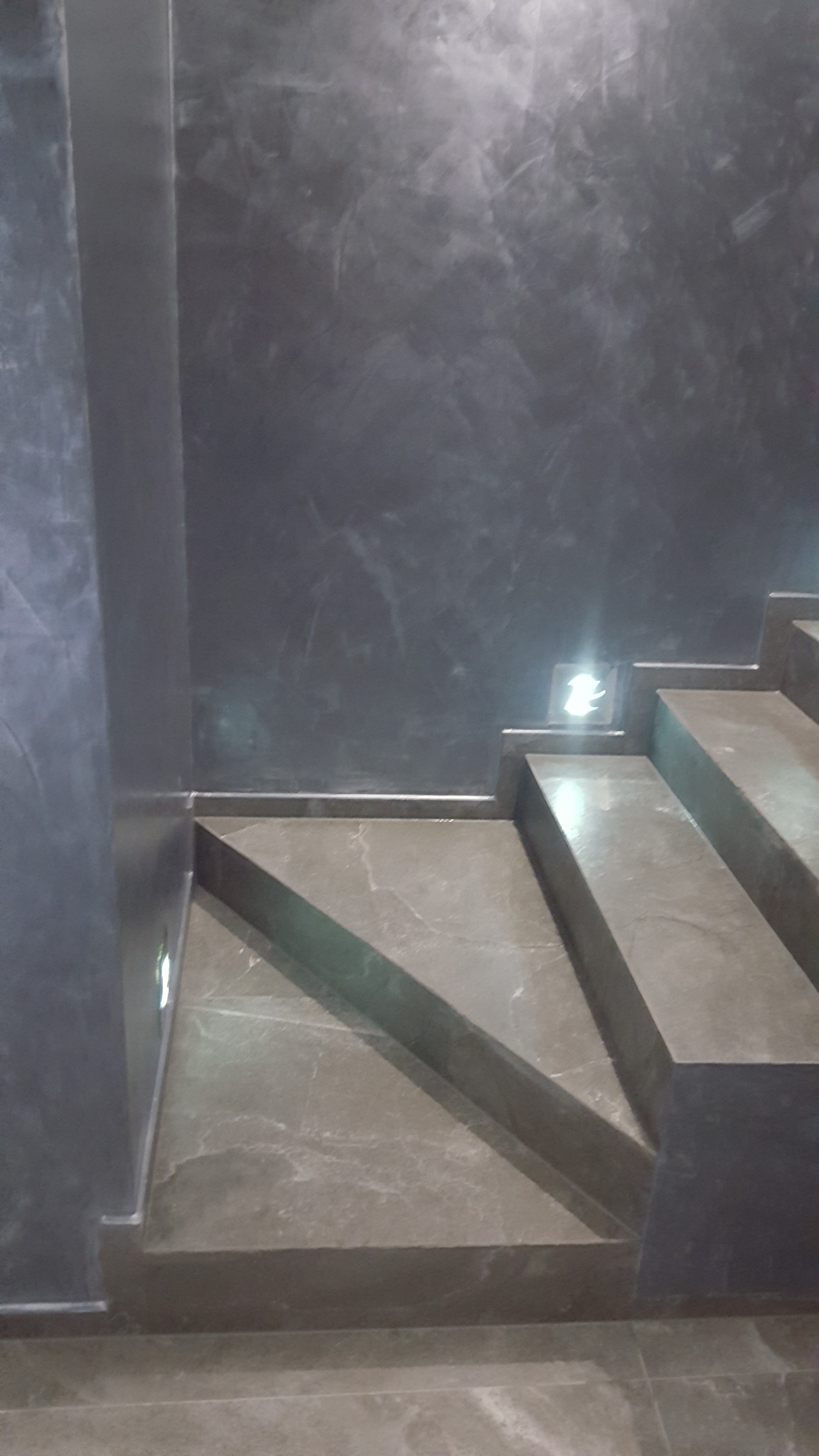 Schody wykończone szarym gresem, ściana pokryta dekoracyjną szarą farbą ze smugami, małe oświetlenie LED na ścianie przy schodach.