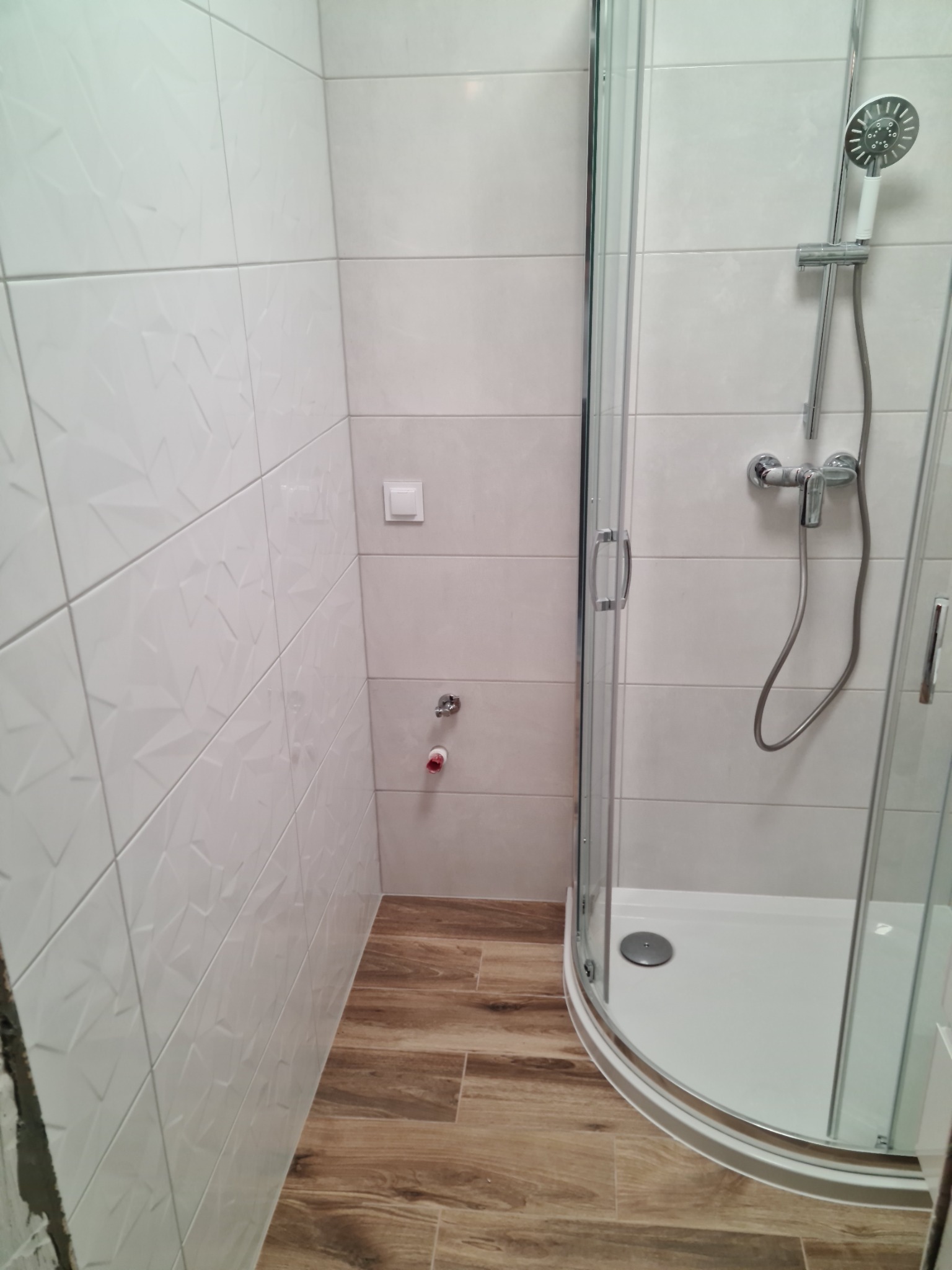 Nowoczesna kabina prysznicowa narożna z chromowaną armaturą i białym brodzikiem, wykończona białymi płytkami z geometrycznym wzorem na ścianie i drewnopodobnymi płytkami na podłodze. Widoczne...