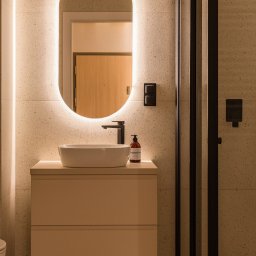zboinskadesign - Elegancka łazienka z owalnym, podświetlanym lustrem nad białą umywalką nablatową. Beżowe płytki, czarna armatura i kabina prysznicowa tworzą spójne, nowoczesne wnętrze.