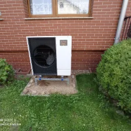 Pompa ciepła Mitsubishi Zubadan zamontowana na aluminiowej konstrukcji na żwirowym podłożu przed ceglaną ścianą domu z oknem.