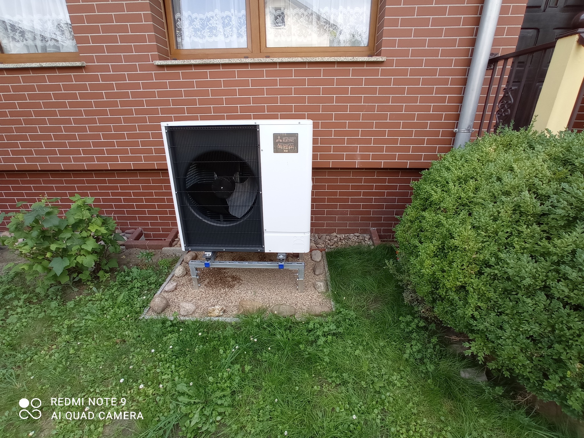 Pompa ciepła Mitsubishi Zubadan zamontowana na aluminiowej konstrukcji na żwirowym podłożu przed ceglaną ścianą domu z oknem.