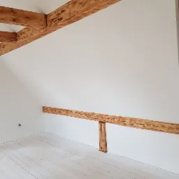 Białe poddasze ze skośnymi ścianami, z widocznymi drewnianymi belkami stropowymi i podłogą pomalowaną na biało, podkreślającymi surowy charakter wnętrza.