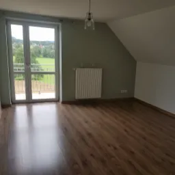 Poddasze po remoncie z drewnianą podłogą, grzejnikiem i wyjściem na balkon z widokiem na zieleń.