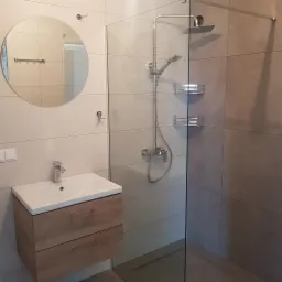 Nowoczesna łazienka z kabiną prysznicową typu walk-in, umywalką nablatową z drewnianą szafką, okrągłym lustrem i beżowymi płytkami w formacie prostokątnym.