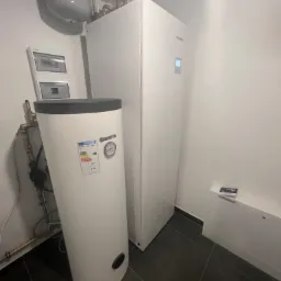 Biała pompa ciepła marki Kaisai i zasobnik ciepłej wody Kospel SE-140 z widoczną etykietą energetyczną, zainstalowane w pomieszczeniu z ciemną podłogą.