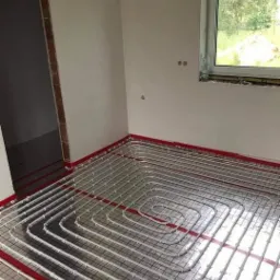 Białe rurki ogrzewania podłogowego ułożone w pętle na czarnej siatce, widoczne czerwone listwy mocujące przy ścianach w surowym wnętrzu z oknem.