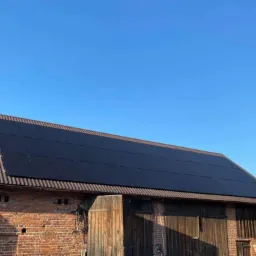 Panel słoneczny na dachu starego, ceglanego budynku gospodarczego w słoneczny dzień. Widoczna brama wjazdowa do budynku.