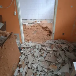 Stos potłuczonych płytek na podłodze w pomieszczeniu z pomarańczowymi ścianami, widoczne ślady kleju i resztki płytek w sąsiednim pomieszczeniu z białymi płytkami, etap demontażu podczas prac...