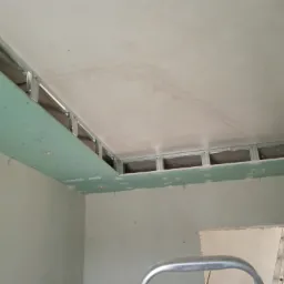 Montaż podwieszanego sufitu z płyt gipsowo-kartonowych na metalowym stelażu, widoczne otwory na oświetlenie punktowe i fragment drabiny w tle.