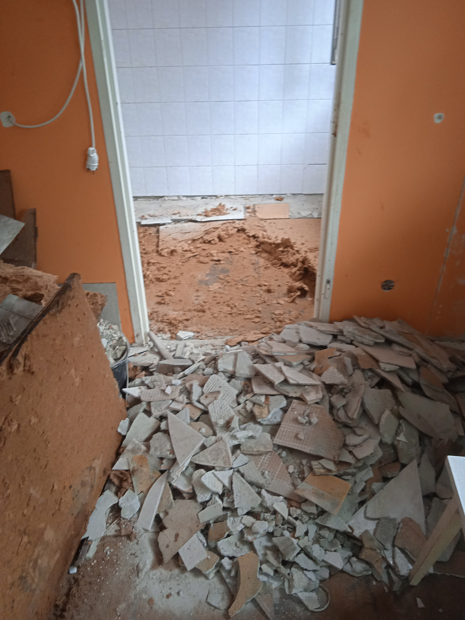 Stos potłuczonych płytek na podłodze w pomieszczeniu z pomarańczowymi ścianami, widoczne ślady kleju i resztki płytek w sąsiednim pomieszczeniu z białymi płytkami, etap demontażu podczas prac...