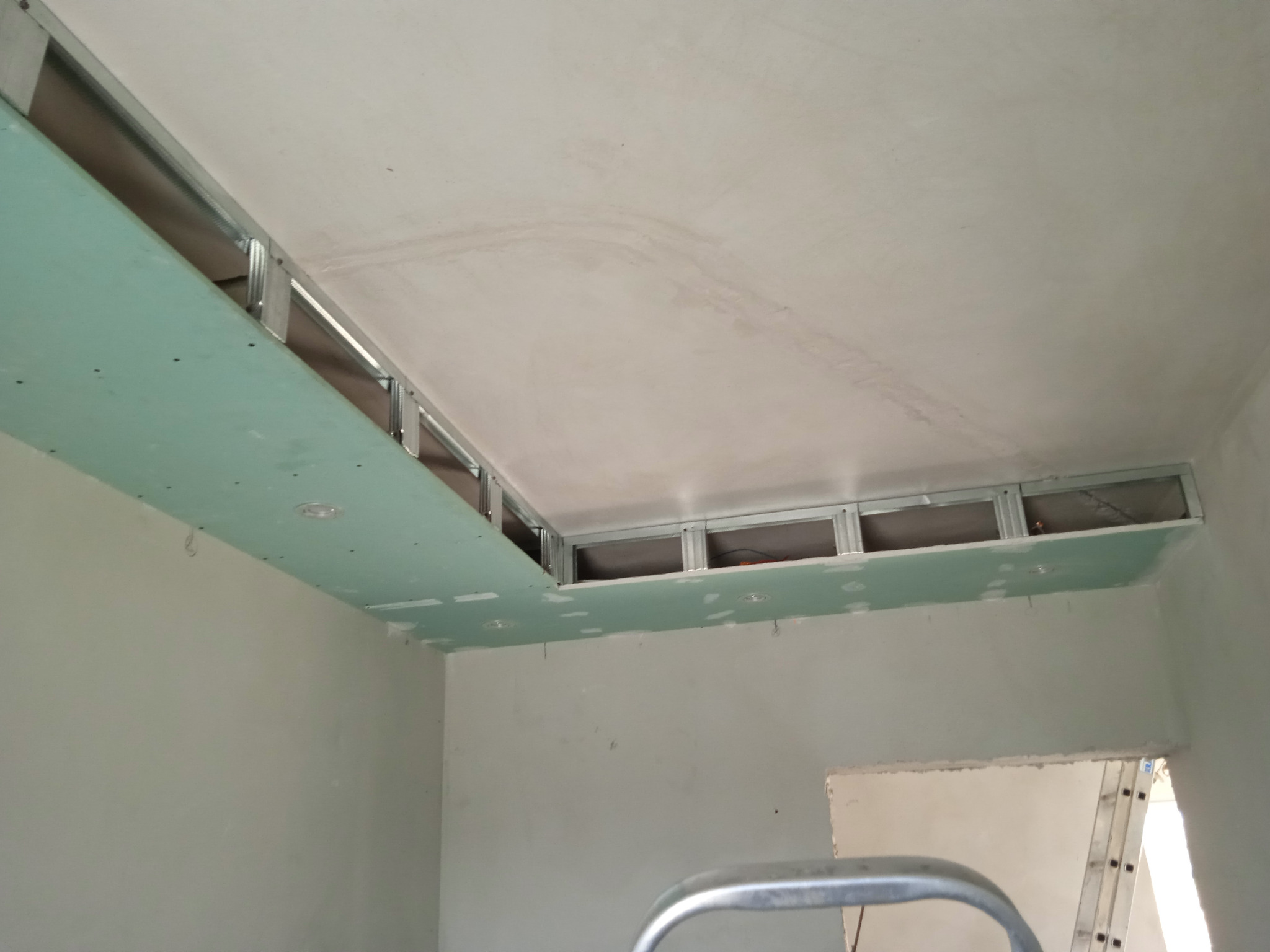 Montaż podwieszanego sufitu z płyt gipsowo-kartonowych na metalowym stelażu, widoczne otwory na oświetlenie punktowe i fragment drabiny w tle.