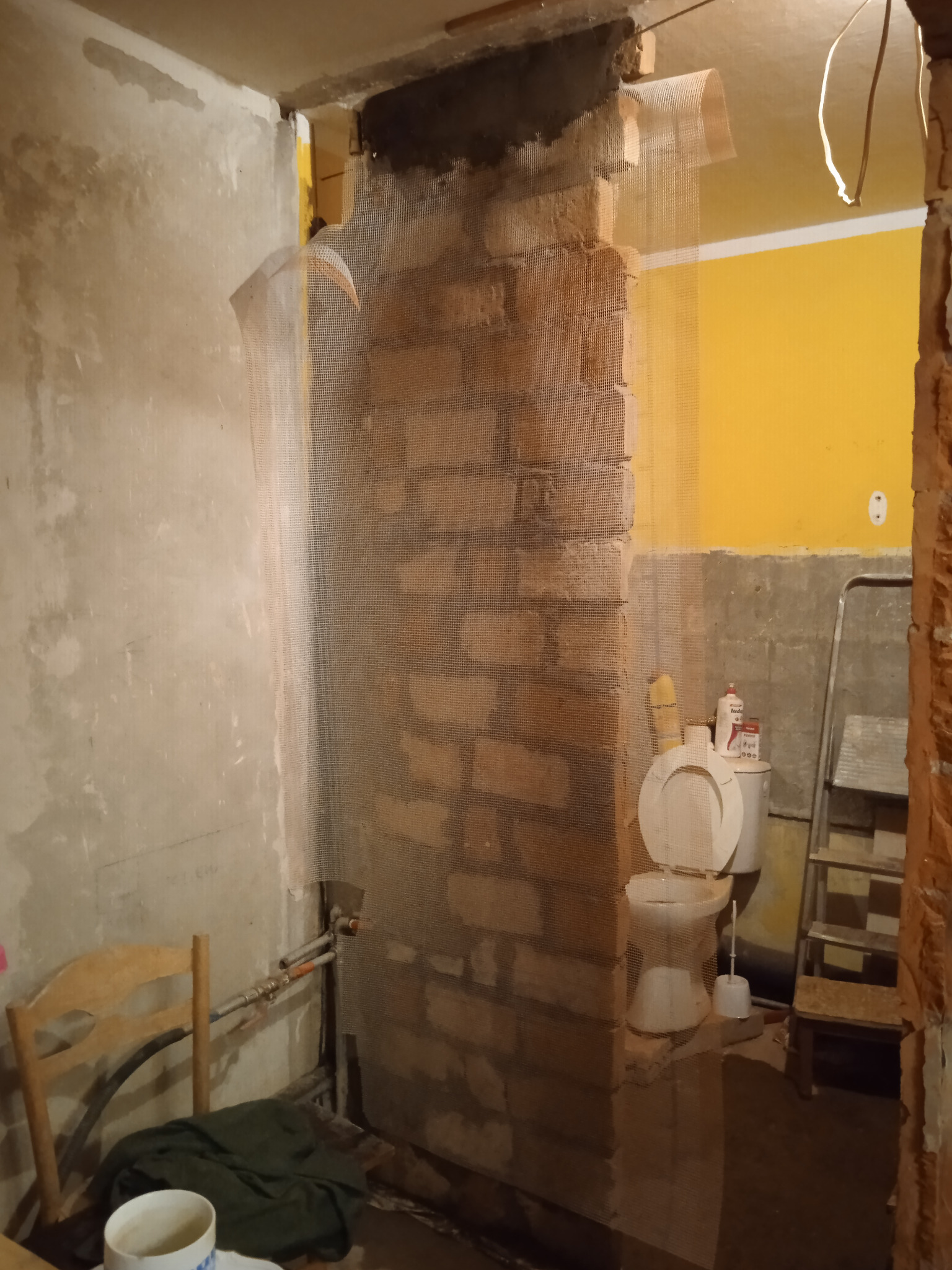 Ceglana ściana wzmocniona siatką pod tynk w trakcie remontu łazienki, widoczna toaleta i drabina, ściany w stanie surowym.