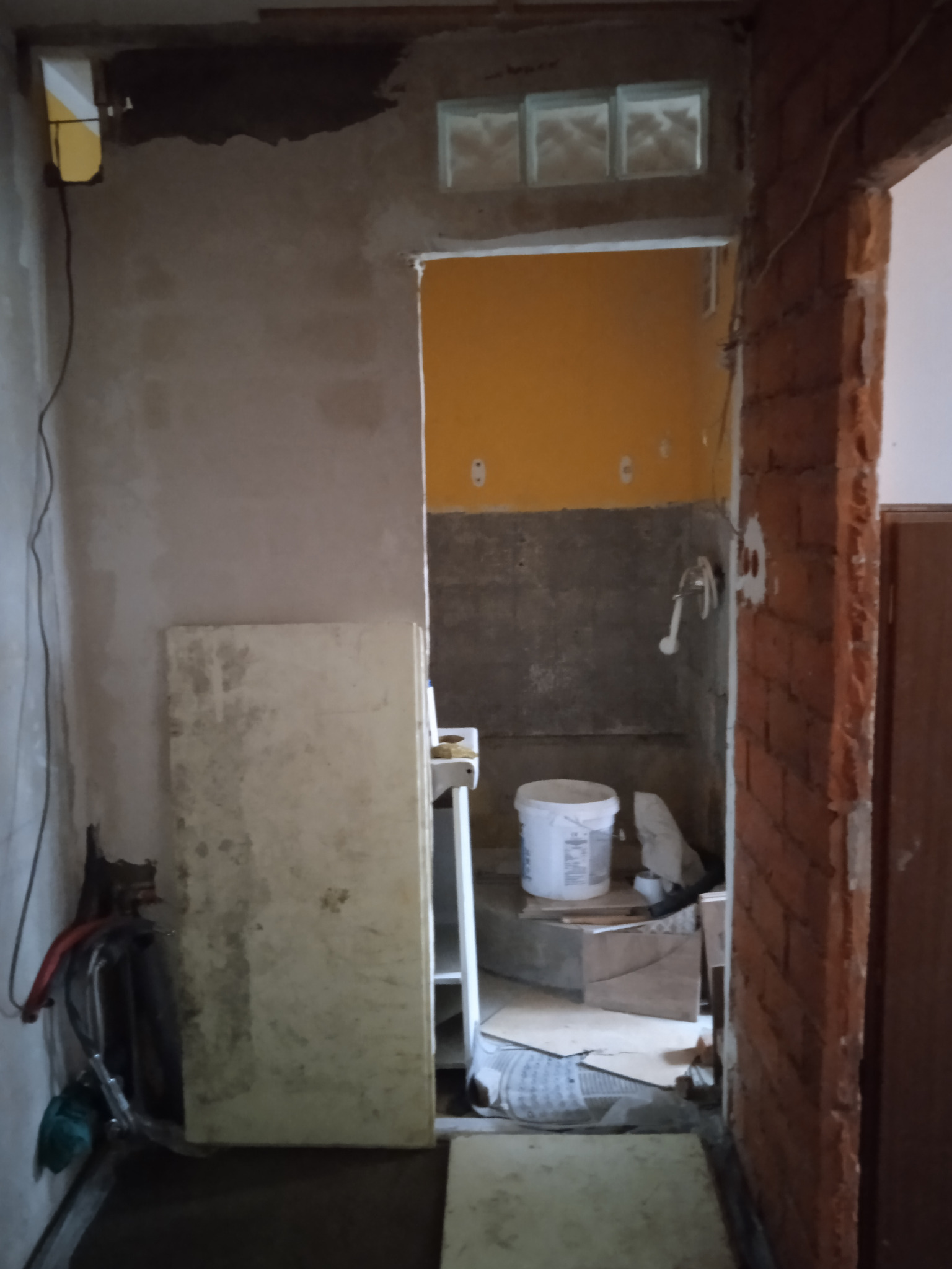 Wnętrze łazienki w trakcie remontu: widoczne surowe ściany, częściowo położone płytki, wiadro z zaprawą, odsłonięta cegła i prowizoryczne zabezpieczenia.