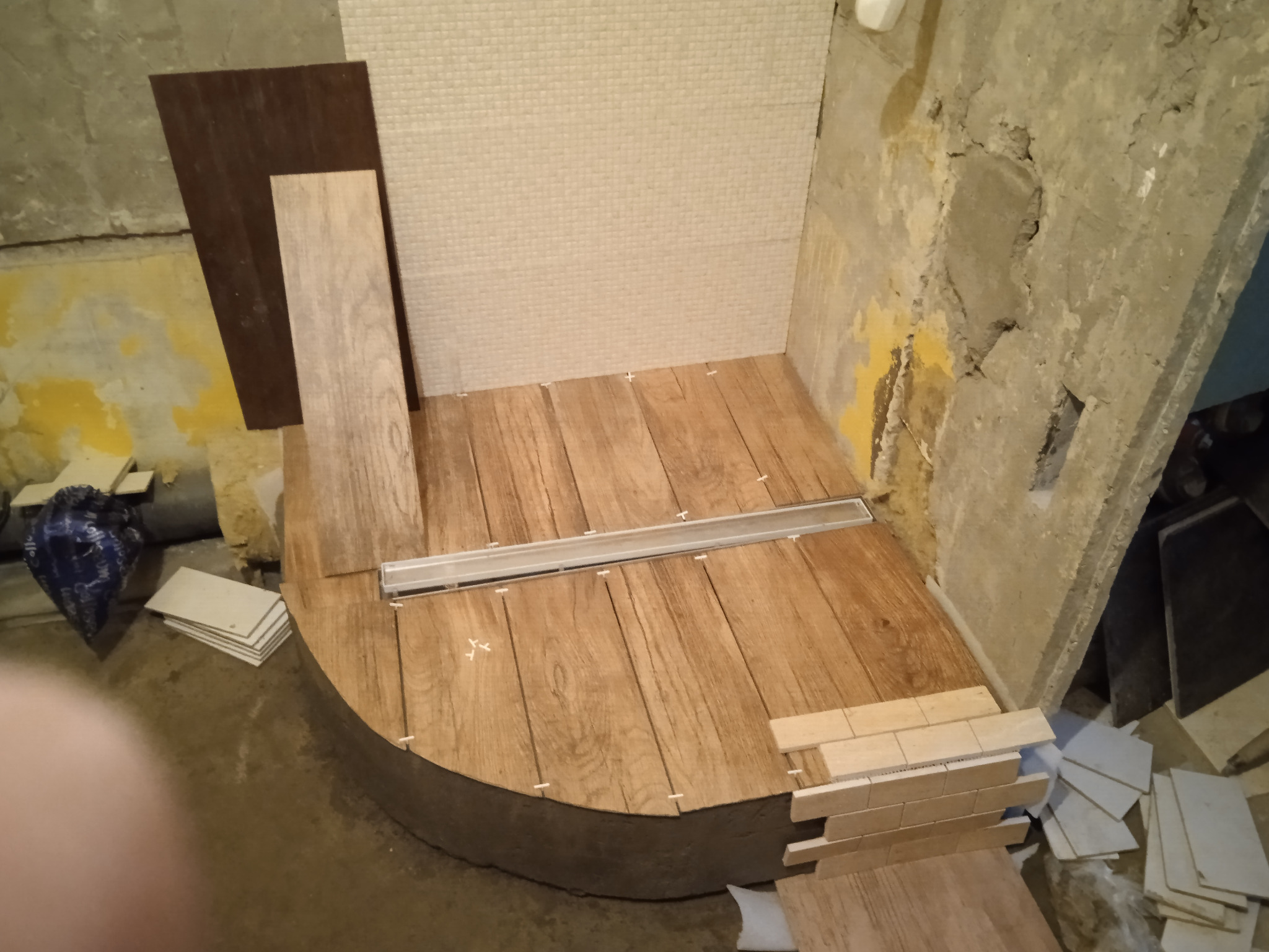 Niedokończona kabina prysznicowa z drewnopodobnymi płytkami na podłodze i jasną mozaiką na ścianie, widoczny odpływ liniowy, zapas płytek i surowy stan ścian w tle.