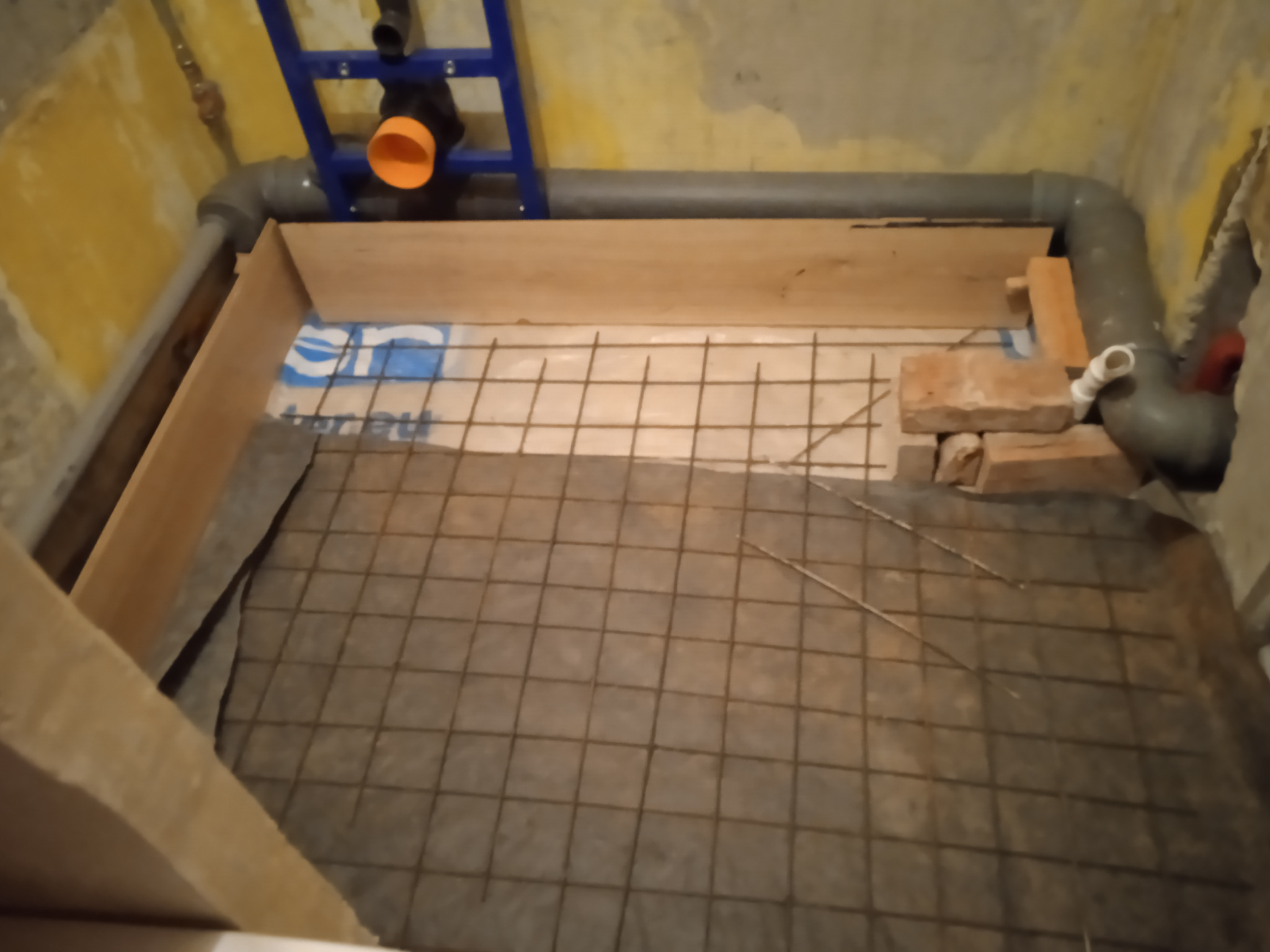 Przygotowanie podłoża pod wylewkę w łazience: widoczna siatka zbrojeniowa na folii izolacyjnej, fragment rur kanalizacyjnych i drewnianego szalunku.