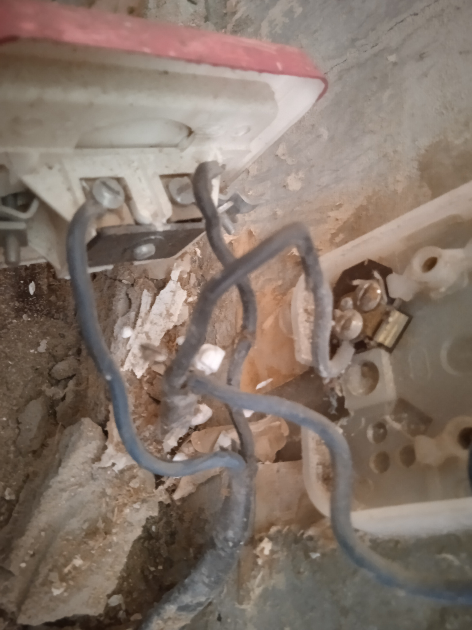 Odsłonięte przewody elektryczne w puszce podtynkowej, widoczne ślady po kuciu w ścianie, fragment starego gniazdka z elementami metalowymi.