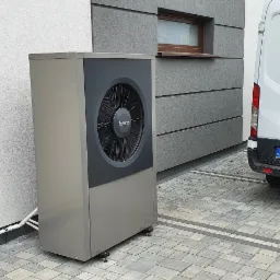 Zewnętrzna jednostka pompy ciepła Buderus zamontowana przy ścianie budynku, obok zaparkowany biały samochód dostawczy Ford Transit z polską tablicą rejestracyjną.