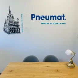 Ściana biura z grafiką przedstawiającą Ratusz w Poznaniu, logo firmy Pneumat oraz hasłem 'Mocni w działaniu'. Na pierwszym planie drewniany stół i dwa szare krzesła, oświetlone lampką.
