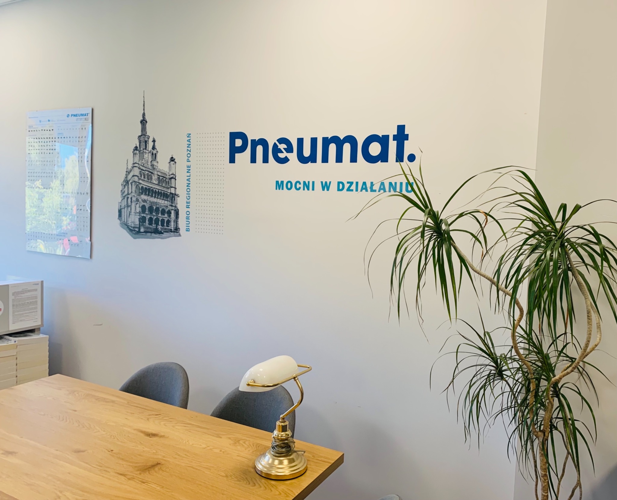 Wnętrze biura z logo firmy Pneumat na ścianie, obok rysunek Starego Rynku w Poznaniu, kalendarz firmowy, drewniany stół, lampka biurkowa i roślina doniczkowa.