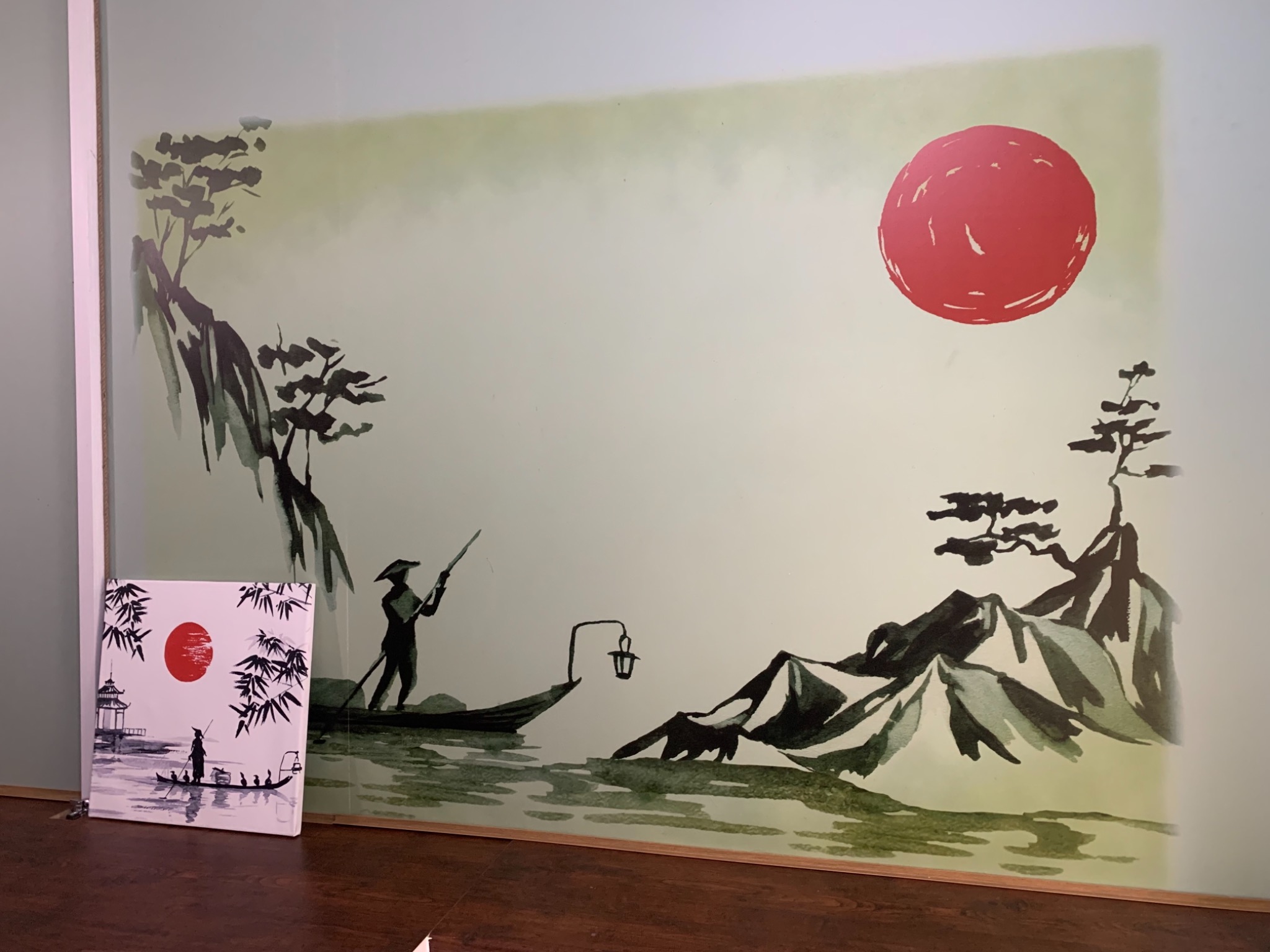 Japoński pejzaż namalowany na ścianie z motywem łodzi, postaci i czerwonego słońca, obok stoi obraz z podobnym motywem.