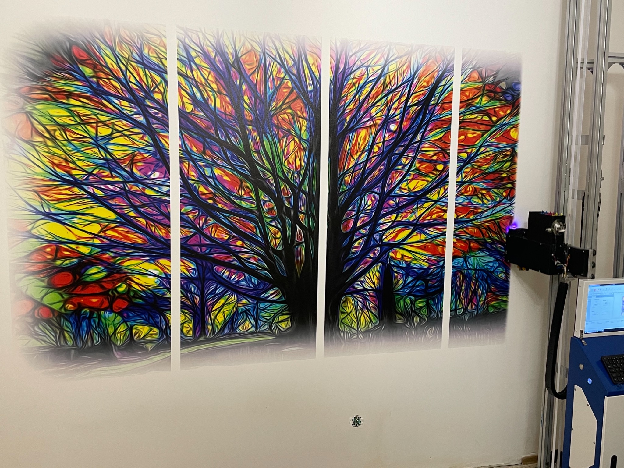 Wieloczęściowy obraz drzewa o intensywnych, tęczowych barwach, wykonany za pomocą drukarki ściennej; widoczny fragment urządzenia drukującego z monitorem i klawiaturą.