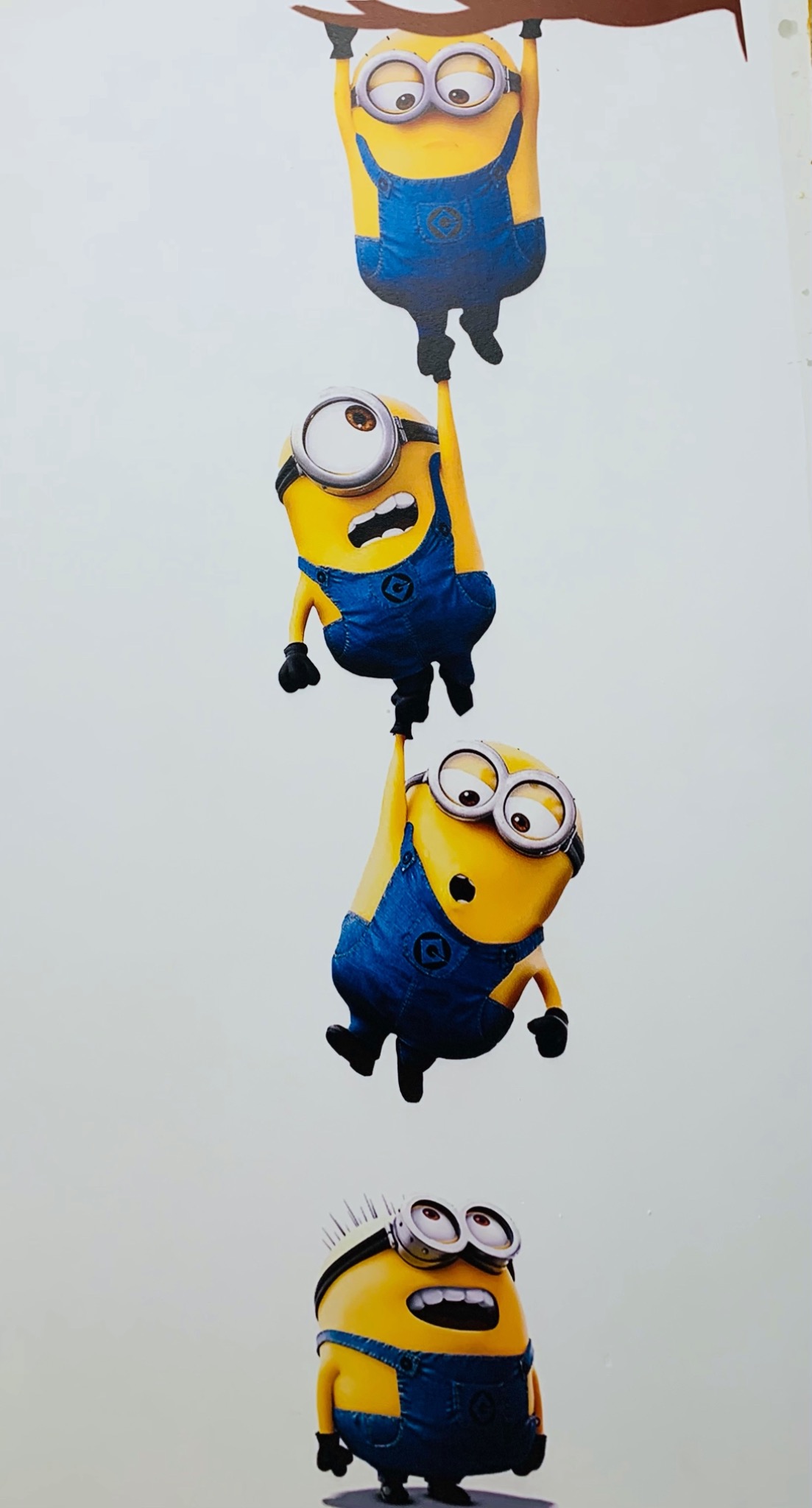 Plakat z czterema Minionkami w kombinezonach, jeden zwisa z gałęzi, dwa trzymają się siebie nawzajem, a ostatni stoi na dole.