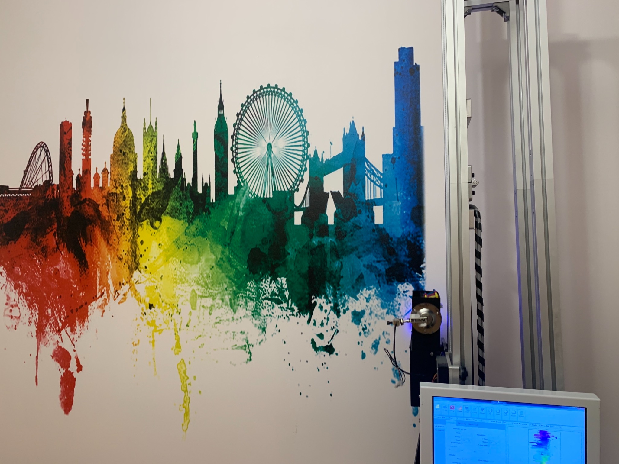 Wielobarwna panorama Londynu, namalowana na ścianie, z widocznym fragmentem maszyny drukującej i ekranem komputera w tle.